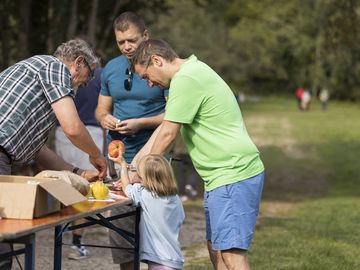 Drei Erwachsene helfen einem kleinen Kind beim Schälen eines Apfels mit Hilfe einer Kurbel an einem Tisch auf einer Wiese.