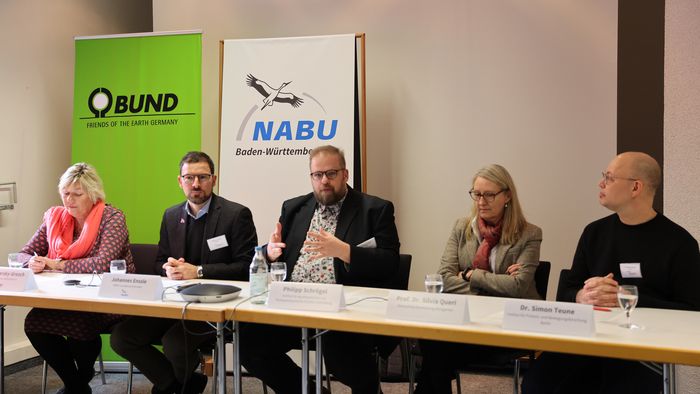 Fünf Menschen sitzen nebeneinander an einem langen Tisch, ener, der in der Mitte sitzt, spricht gerade. Im Hintergrund zwei Hochkant-Banner mit Schriftzügen BUND und NABU Baden-Württemberg.