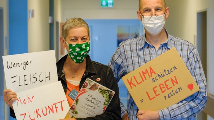 Ein Mann und eine Frau stehen in einem Krankenhausflur und halten Schilder mit Forderungen für mehr Klimaschutz in den Händen.