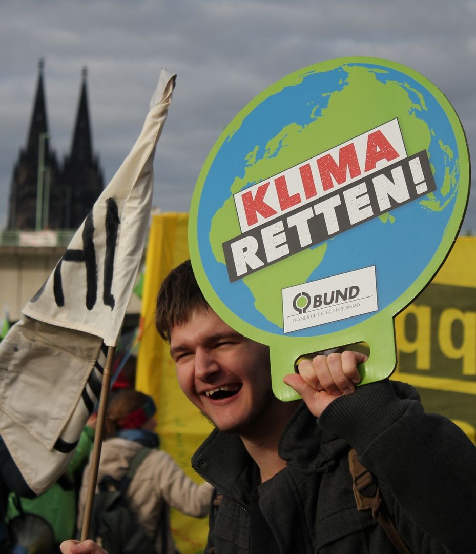 Ein Junger Mann hält ein Schild mit einer Erde und dem Schriftzug "Klima retten" in die Höhe. Links im Hintergrund erkennt man den Kölner Dom.