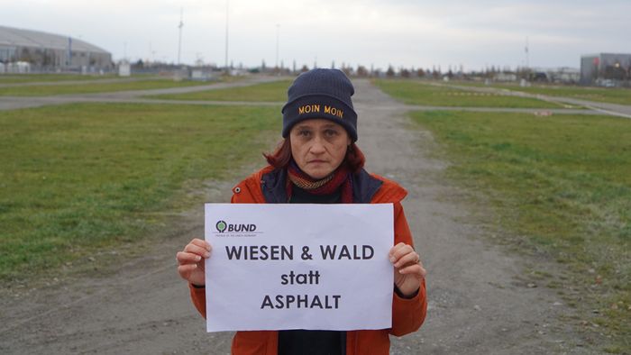 Eine Frau mit roter Jacke steht vor dem Gelände der MEsse Karlsruhe und hat ein Schild mit der Aufschrift "Wiesen und Wald. statt Asphalt" in den Händen.