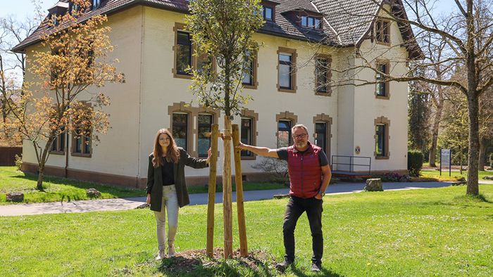 Klimamanagerin Sophia Lager und der Leiter der Anlagenpflege, Martin Theobold, stehen neben einer jungen Steineiche (Quercus ilex) im Park des Klinikums am Weissenhof.
