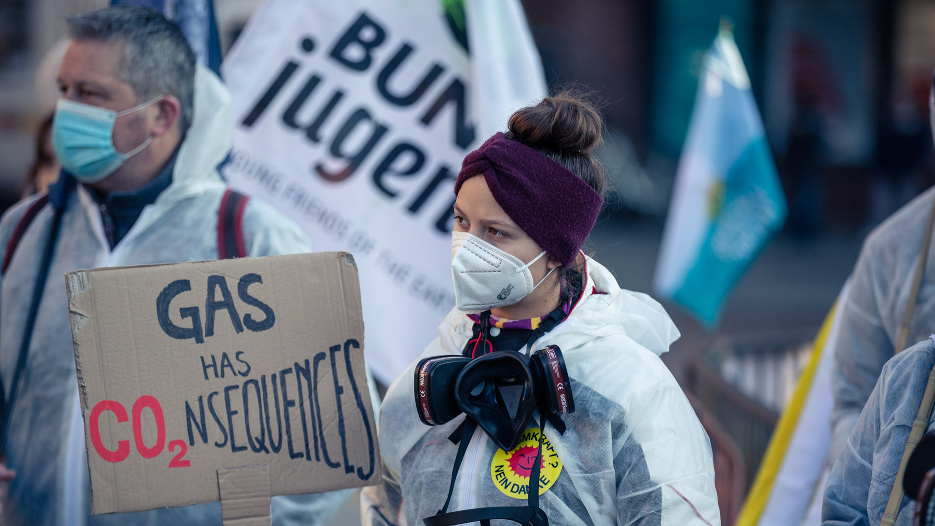 Eine junge Frau im weißen Schutzanzug hält ein Schild, auf dem "Gas has Consequences" steht.