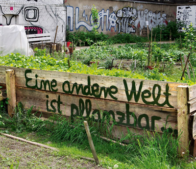 Ein verwilderter Garten mit einem mit Latten umzäumtes Beet auf dem steht "Eine andere Welt ist pflanzbar". 